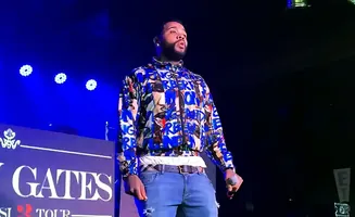 A man wearing a colorful jacket and jeans stands on stage under blue lights, holding a microphone.