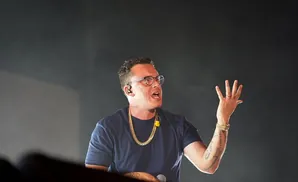 Rapper Logic performs on stage, wearing glasses, a gold chain, and a dark blue t-shirt, holding a microphone.