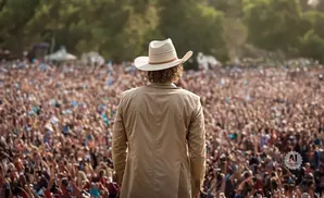 Man in a cowboy hat and tan jacket facing a cheering crowd at an outdoor concert.