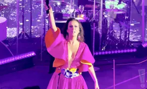 A woman in a pink and orange dress sings into a microphone on a stage with purple lights.