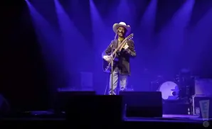 A man in a cowboy hat plays an acoustic guitar on a dimly lit stage, with blue spotlights overhead.