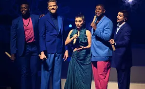 A diverse group of five people in bright blue suits and a woman in a teal dress stand on a stage with a snowflake backdrop.