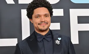 Trevor Noah smiles at the camera wearing a dark suit, bow tie, and a star brooch with a blue heart pin.