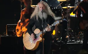Musician with long white hair and beard plays a guitar covered in signatures while on stage.