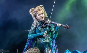 Violinist with blonde hair and a headdress plays on a stage with red lighting.
