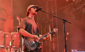 A male guitarist in a cap and t-shirt sings into a microphone while playing a dark electric guitar on stage.