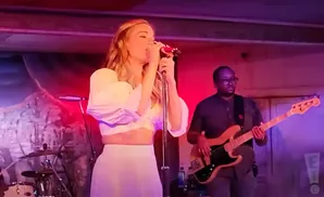 Singer in white crop top and skirt performs on stage with a bassist and drummer.