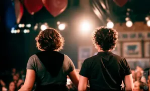 Two people with curly hair stand with their backs to the camera, facing a bright spotlight on a stage.