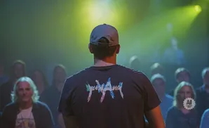 Man in a cap and t-shirt with a colorful design faces away from the camera, with an audience behind him lit by yellow spotlights.