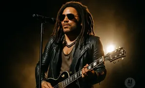 Lenny Kravitz plays guitar and sings into a microphone on a dimly lit stage.