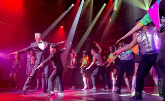 Dancers in sparkling outfits perform on a stage with red and white spotlights.