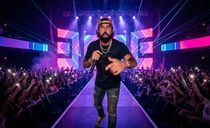 A male performer stands on stage singing into a microphone to a cheering crowd holding up cell phones.