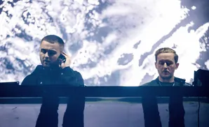 Two men stand behind a DJ booth with a large, abstract image projected behind them.
