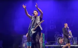 A man with a guitar sings into a microphone on a stage lit by blue lights, while another man plays a trombone.