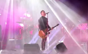 A male singer with tattoos plays a guitar on a smoke-filled stage with pink and white lights.