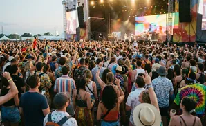 Woman in a floral dress and hat at an outdoor festival with colorful tents and blurred people.