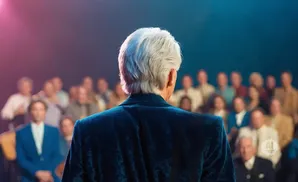 A person with white hair in a blue jacket speaks to an audience.