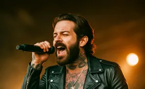 Man with beard and tattoos singing into a microphone, wearing a leather jacket.