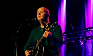 A bald man in a dark green sweater plays an acoustic guitar on stage with purple lights in the background.
