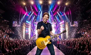 Musician plays a gold electric guitar on stage with confetti and fireworks in front of a cheering crowd.