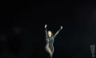 A woman in a sparkly black dress with sheer panels sings with her arms raised in a concert.