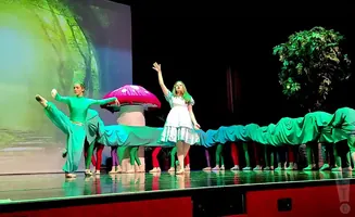 Dancers in a play perform on stage, with one dressed as a caterpillar and others as its segments, against a forest backdrop.