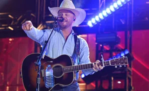 A man in a cowboy hat sings and plays an acoustic guitar on stage, illuminated by blue and red lights.