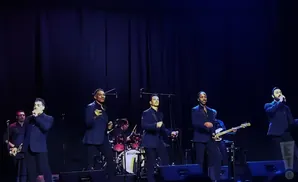 Five men in suits perform on a stage with a dark blue backdrop.