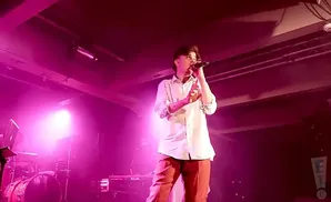 A male singer in a white shirt and orange pants performs on a dimly lit stage with pink lighting.