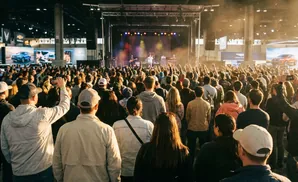 A crowd of people watches a band perform on stage at a bright, indoor event.