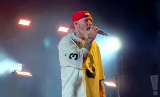 A tattooed man in a red baseball cap and a white and yellow shirt sings into a microphone on a stage with bright lights.