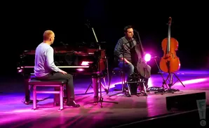 Musicians play piano and cello on a stage with purple lighting.