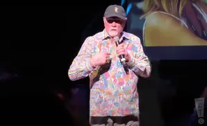 A man wearing a colorful, floral-patterned shirt and a baseball cap holds a microphone on stage.