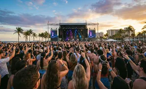 A large crowd at an outdoor beach concert enjoys a band performing on a stage at sunset, with palm trees and buildings in the background.