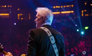 A man with white hair plays a guitar on a stage with a crowd in the background.
