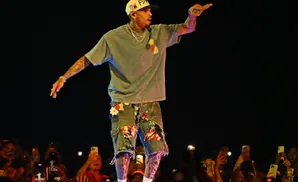 Chris Brown performs on stage, wearing a grey t-shirt, distressed denim shorts, and a baseball cap, with a crowd holding up phones in the foreground.
