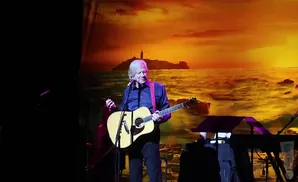 A man plays an acoustic guitar on stage in front of a sunset ocean backdrop.