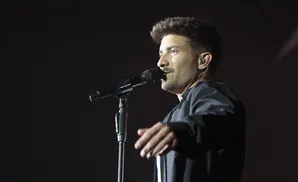 A man with dark hair sings into a microphone on stage.
