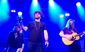 Three musicians performing on a stage with blue spotlights: a guitarist on the right, a singer in the center with a hat and necklace, and another musici…