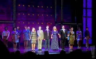 A diverse cast of performers in stylish outfits take a bow on a stage with purple lighting and sparkly accents.