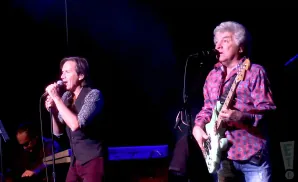 Two male musicians perform on stage, one singing into a microphone, the other playing a bass guitar.