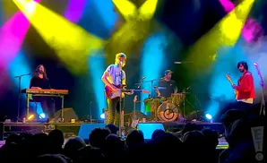 A band performs on a stage with bright yellow and pink lights and fog.