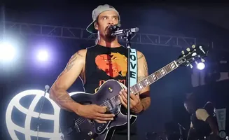 Musician with tattoos, wearing a baseball cap and a tank top with a heart design, plays an electric guitar on stage.