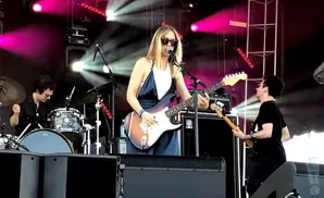 A band performs on a stage with pink and white lights. A woman sings and plays guitar while a drummer and bassist play their instruments.