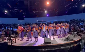 A band in matching purple and orange outfits performs on stage with trumpets and saxophones in front of a large audience.