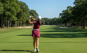 A female golfer in a maroon skirt and shirt swings her club on a golf course with trees and sand traps.