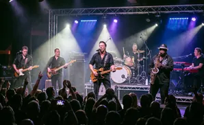 A band performs on stage with a saxophonist, guitarist, drummer, and keyboardist, lit by purple and white spotlights.