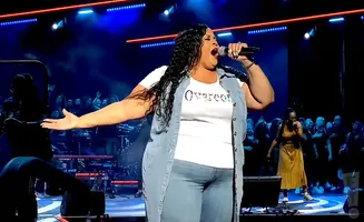 A woman sings into a microphone on stage with her arm outstretched.