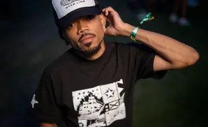 A man with a goatee wears a black t-shirt with a white graphic and a trucker hat.