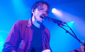 A man with dark hair sings into a microphone on a blue-lit stage, wearing a dark shirt and a maroon jacket.